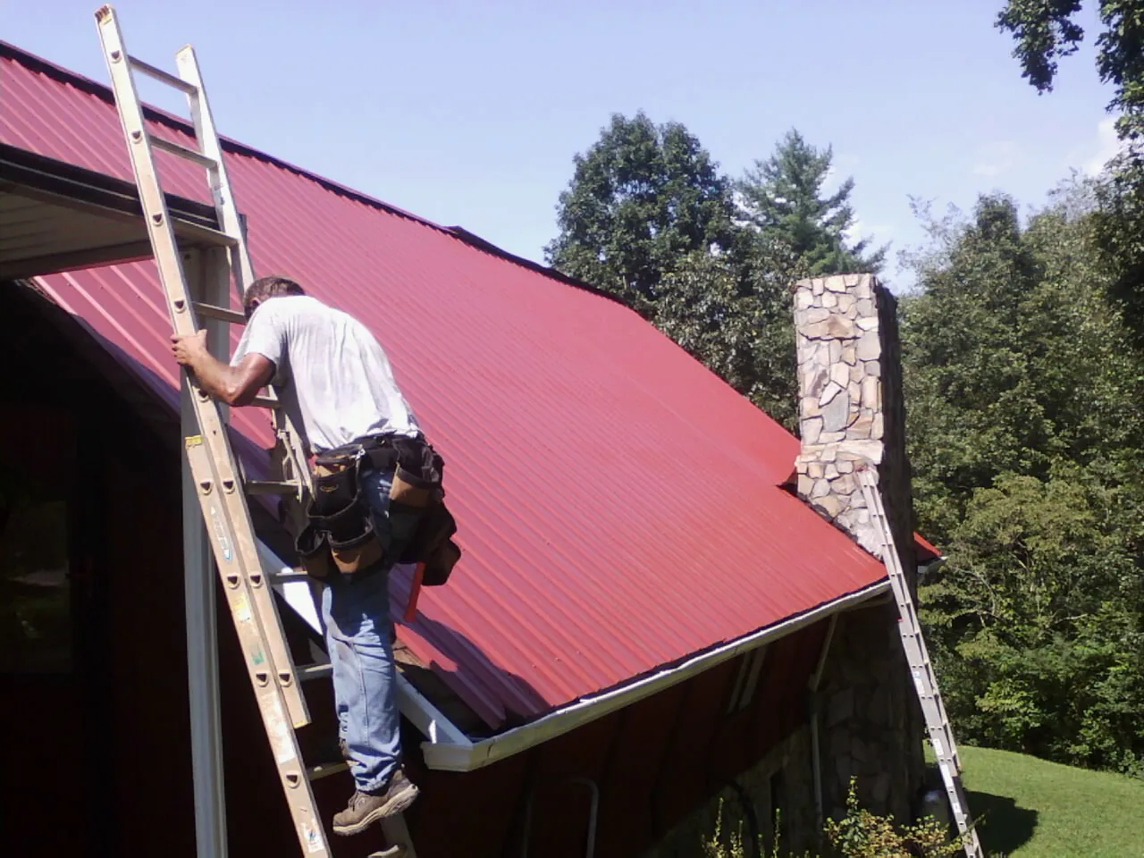 Roofer on a ladder beside a red metal roof and stone chimney — Soffit & Fascia Repair by Roofing Brothers in West Liberty